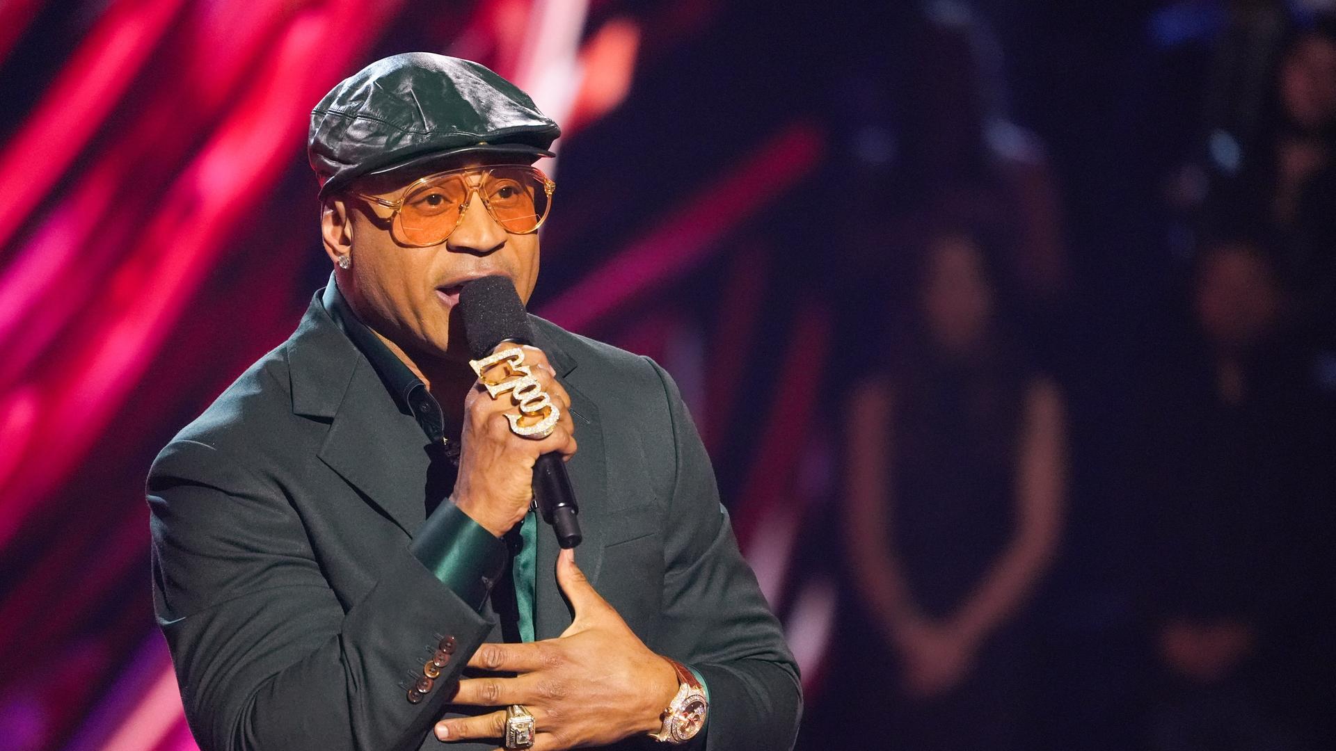 LL Cool J trägt eine Schiebermütze und hat ein Mikrofon in der Hand. Er hat einen Ring, der die Buchstaben "Cool J" formt. LL Cool J trägt eine Schiebermütze und hat ein Mikrofon in der Hand. Er hat einen Ring, der die Buchstaben "Cool J" formt.