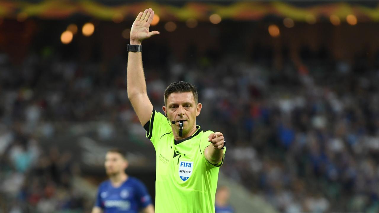 Ein Mann im gelben Schiedsrichter-Trikot und Trillerpfeife im Mund hat den rechten Arm erhoben und den linken Arm ausgestreckt.