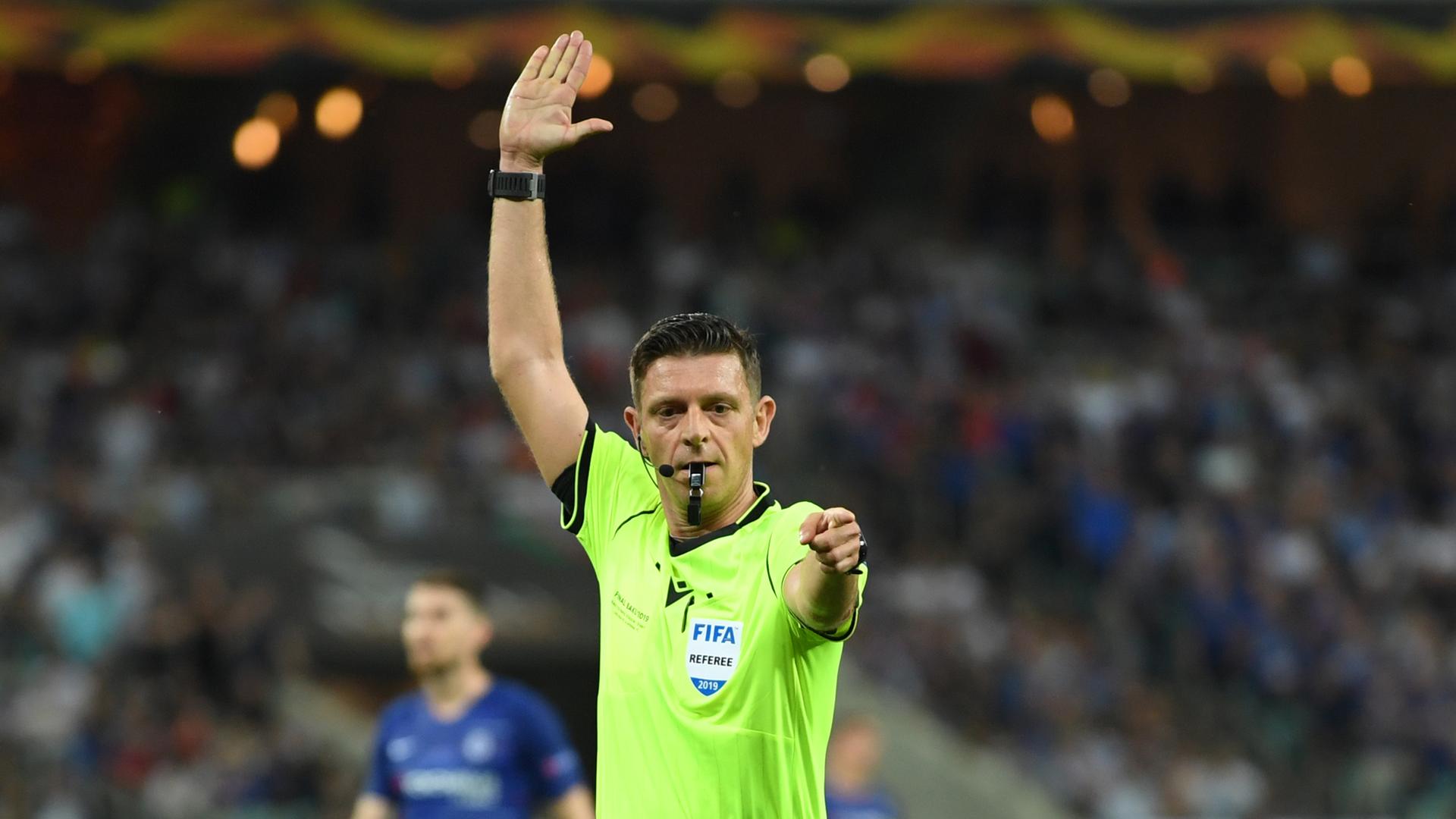 Ein Mann im gelben Schiedsrichter-Trikot und Trillerpfeife im Mund hat den rechten Arm erhoben und den linken Arm ausgestreckt.