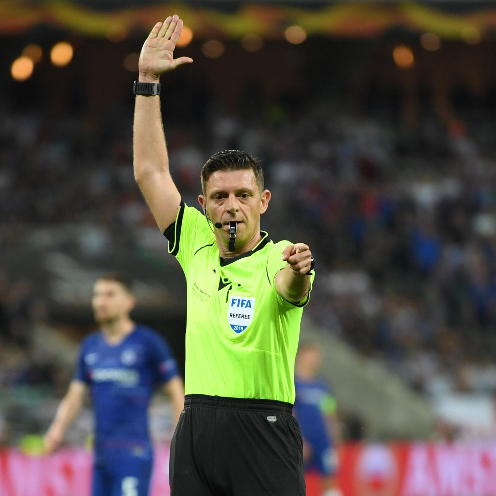 Ein Mann im gelben Schiedsrichter-Trikot und Trillerpfeife im Mund hat den rechten Arm erhoben und den linken Arm ausgestreckt.