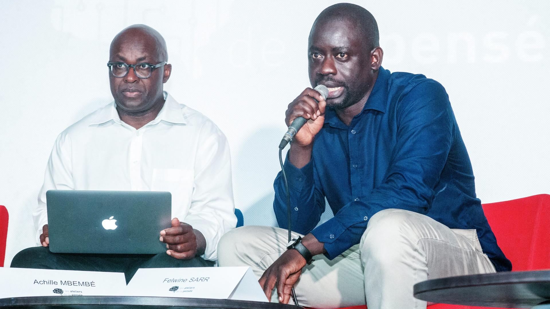 Achille Mbembe und Felwine Sarr sitzen an einem kleinen Tisch. Mbembe hat einen Laptop auf dem Schoß, Sarr spricht in ein Mikrofon.