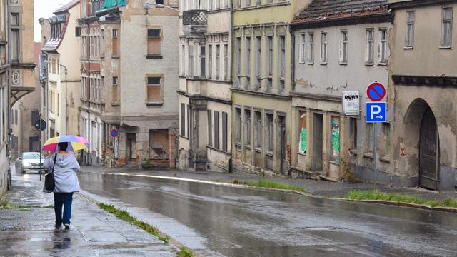 Eine Frau mit buntem Regenschirm läuft bei Regen eine Straße entlang. auf der anderen Straßenseite sieht man einige verfallene bzw. leer stehende Häuser.