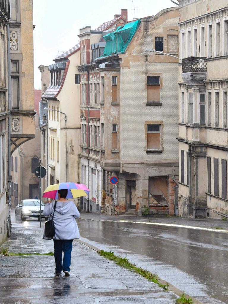 Eine Frau mit buntem Regenschirm läuft bei Regen eine Straße entlang. auf der anderen Straßenseite sieht man einige verfallene bzw. leer stehende Häuser.