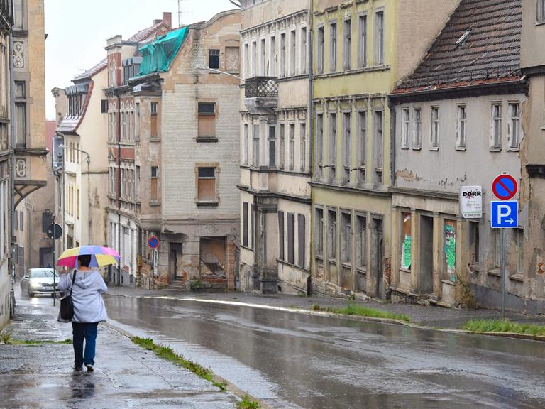 Eine Frau mit buntem Regenschirm läuft bei Regen eine Straße entlang. auf der anderen Straßenseite sieht man einige verfallene bzw. leer stehende Häuser.