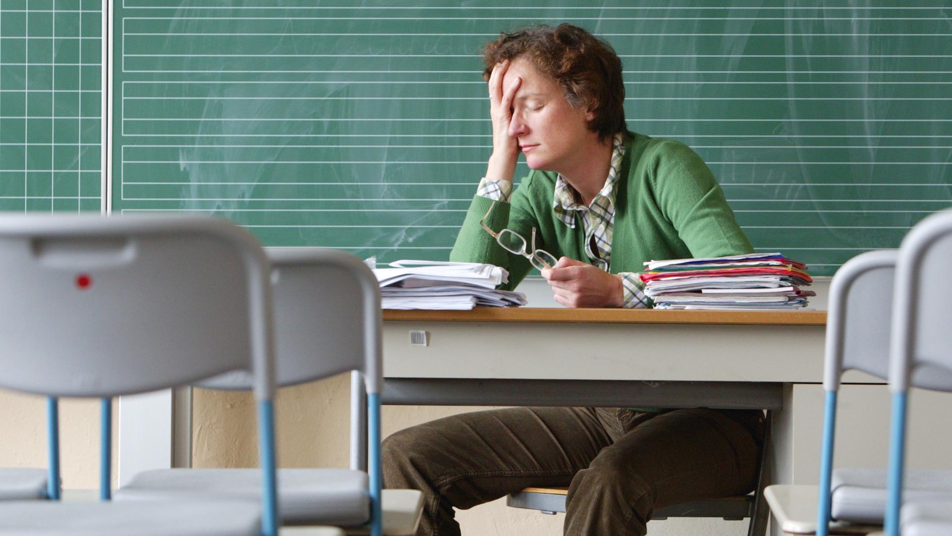Eine Lehrerin sitzt alleine in einem Klassenraum und fasst sich, die Augen geschlossen, an die Stirn.