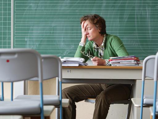Eine Lehrerin sitzt alleine in einem Klassenraum und fasst sich, die Augen geschlossen, an die Stirn.