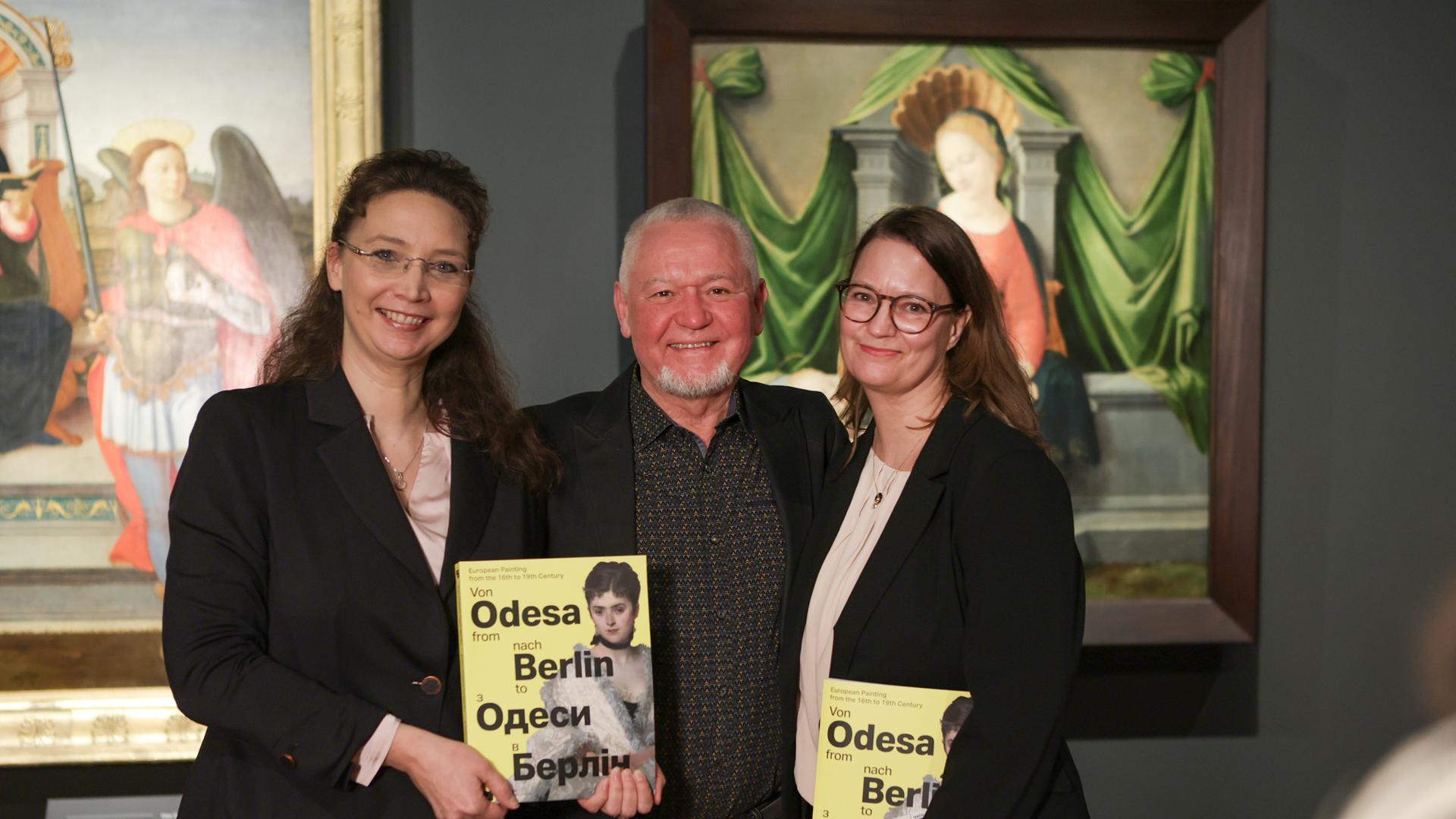 Zwei Frauen und ein Mann in ihrer Mitte präsentieren den Ausstellungskatalog der Schau "Von Odsea nach Berlin. Europäische Malerei des 16. bis 19. Jahrhunderts"). Im Hintergrund sind Bilder in den Ausstellungsräumen zu sehen. 