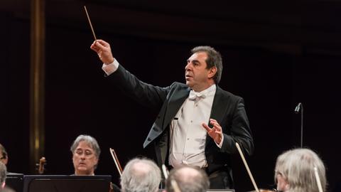 Ein Dirigent hebt seinen rechten Arm mit Taktstock, um dem Orchester etwas anzuzeigen. Vom Orchester sieht man nur ein paar Geiger und Geigerinnen. 