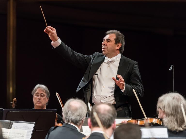 Ein Dirigent hebt seinen rechten Arm mit Taktstock, um dem Orchester etwas anzuzeigen. Vom Orchester sieht man nur ein paar Geiger und Geigerinnen. 