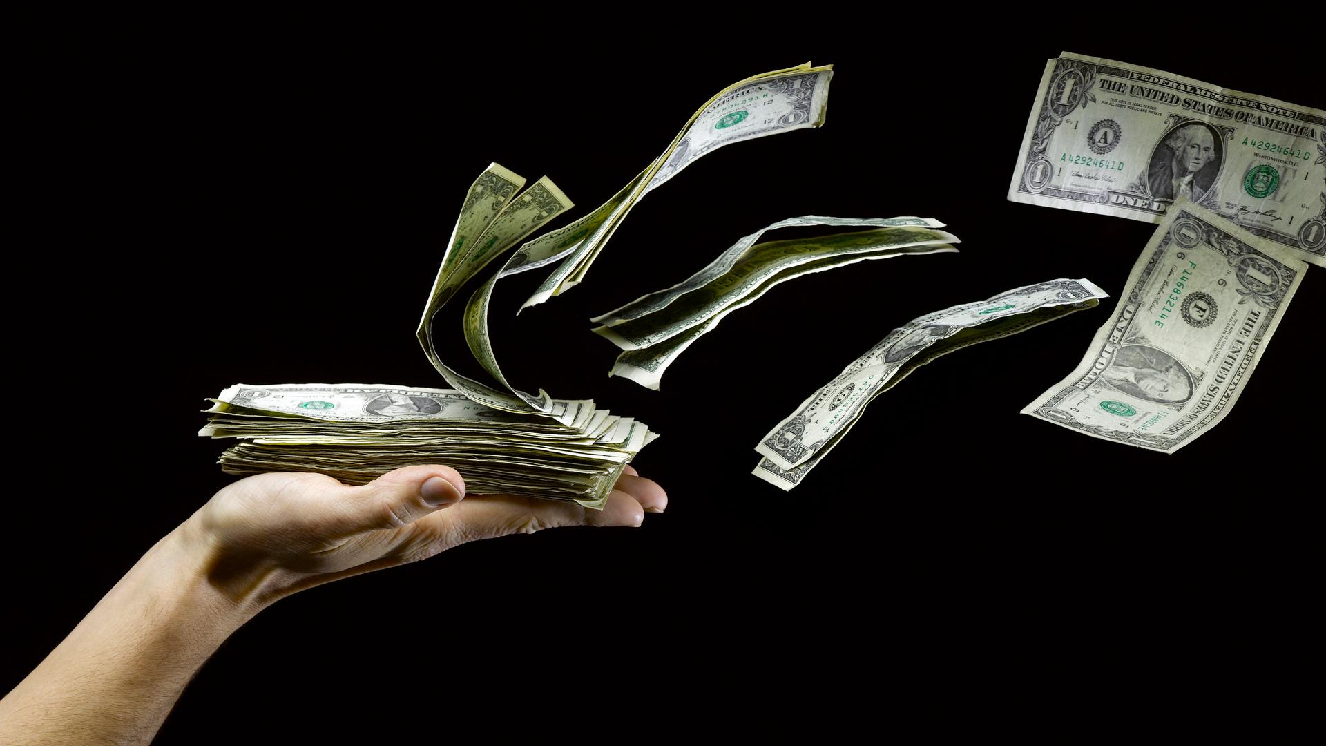 US Dollar fliegen von einer ausgestreckten Hand, die einen Haufen Geldscheine hält.