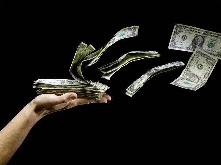 US Dollar fliegen von einer ausgestreckten Hand, die einen Haufen Geldscheine hält.
