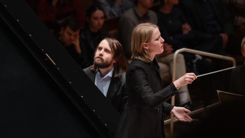 Hinter der Dirigentin Reinhardt ist der aufgeklappte Flügel sichtbar, an dem der Pianist Trifonov sitzt und mit erhobenem Kopf spielt.