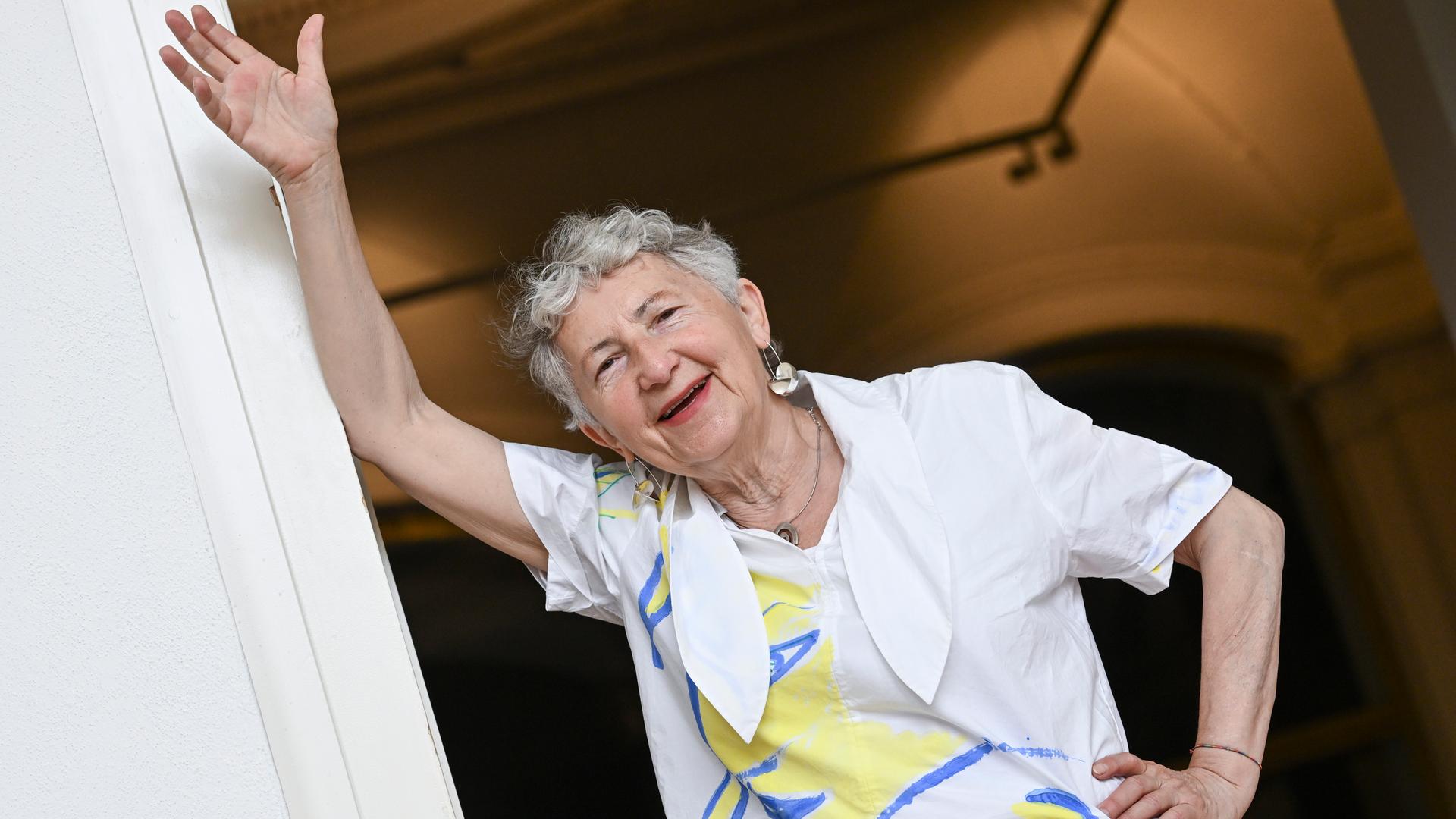 Die Künstlerin Gabriele Stötzer steht lachend an einem Türpfosten in ihrer Ausstellung „Dabei sein und nicht schweigen“ im Gropius Bau. Sie trägt eine weiße Jacke und nimmt eine lässige Pose ein. 