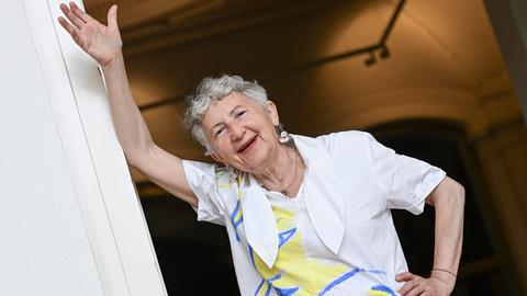 Die Künstlerin Gabriele Stötzer steht lachend an einem Türpfosten in ihrer Ausstellung „Dabei sein und nicht schweigen“ im Gropius Bau. Sie trägt eine weiße Jacke und nimmt eine lässige Pose ein. 