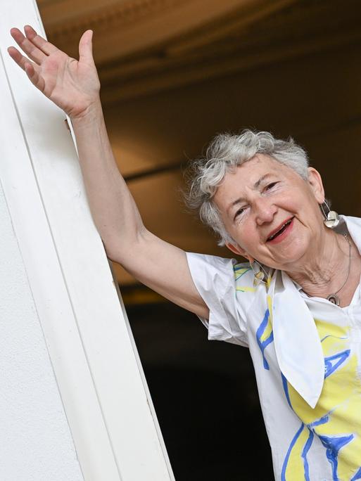 Die Künstlerin Gabriele Stötzer steht lachend an einem Türpfosten in ihrer Ausstellung „Dabei sein und nicht schweigen“ im Gropius Bau. Sie trägt eine weiße Jacke und nimmt eine lässige Pose ein. 