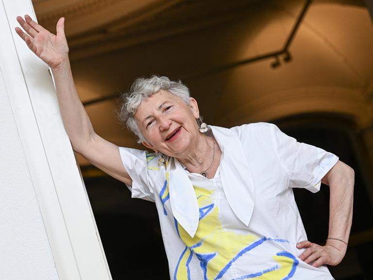 Die Künstlerin Gabriele Stötzer steht lachend an einem Türpfosten in ihrer Ausstellung „Dabei sein und nicht schweigen“ im Gropius Bau. Sie trägt eine weiße Jacke und nimmt eine lässige Pose ein. 
