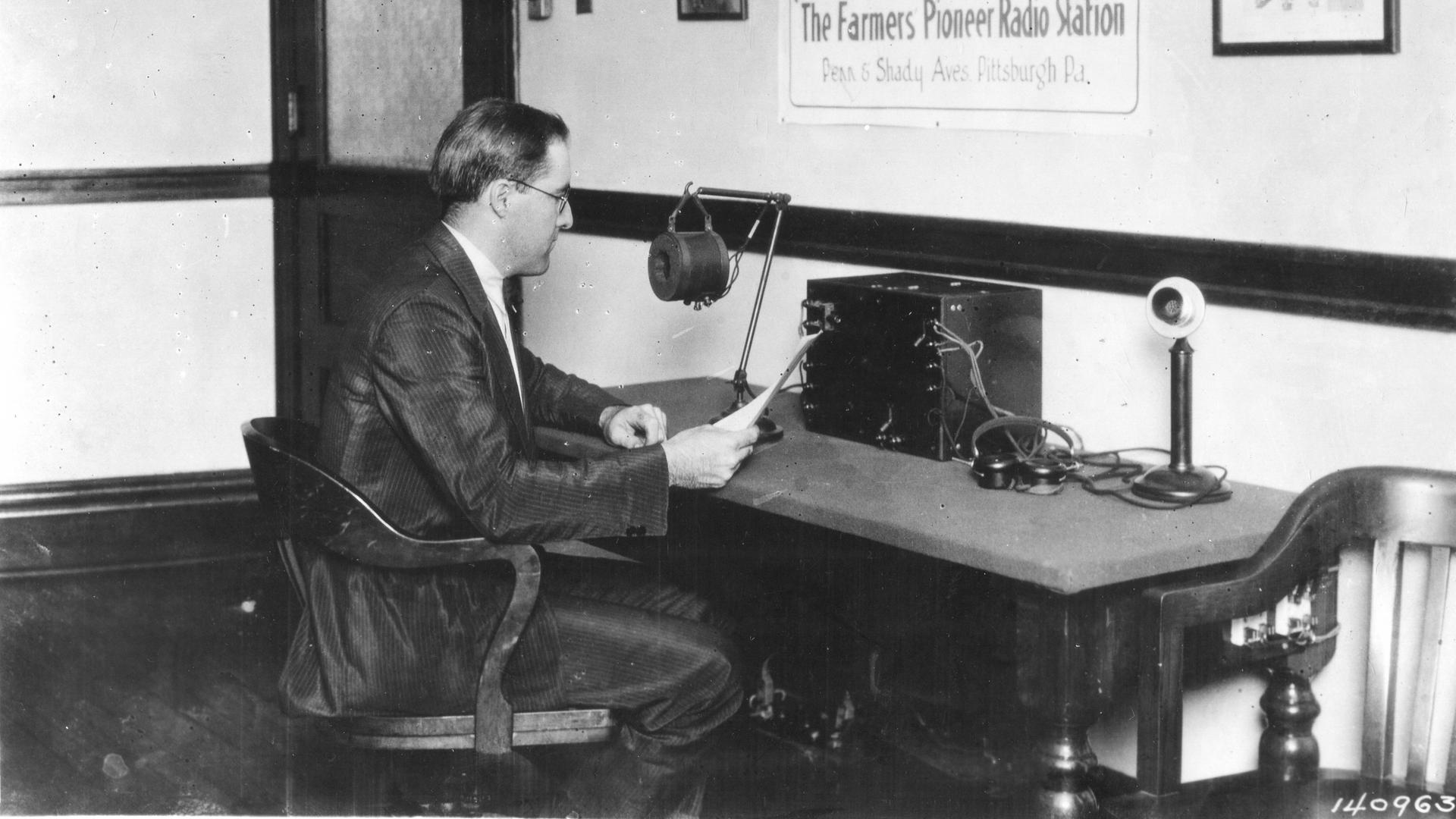 Ein Mann, Frank Mullen, im weißen Hemd und dunklem Anzug sitzt 1922 in einem Stuhl an einem Tisch, auf dem ein Mikrofon steht. Er hält ein Blatt Papier in der Hand und schaut drauf. An der Wand hängt ein Poster mit der Aufschrift KDKA in Großbuchstaben, "The National Stockmans & Farmer Radio Station of KDKA" und "The Farmers Pioneer Radio Show" 