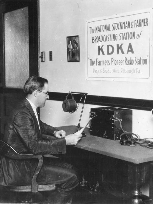 Ein Mann, Frank Mullen, im weißen Hemd und dunklem Anzug sitzt 1922 in einem Stuhl an einem Tisch, auf dem ein Mikrofon steht. Er hält ein Blatt Papier in der Hand und schaut drauf. An der Wand hängt ein Poster mit der Aufschrift KDKA in Großbuchstaben, "The National Stockmans & Farmer Radio Station of KDKA" und "The Farmers Pioneer Radio Show" 