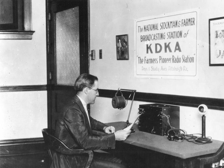 Ein Mann, Frank Mullen, im weißen Hemd und dunklem Anzug sitzt 1922 in einem Stuhl an einem Tisch, auf dem ein Mikrofon steht. Er hält ein Blatt Papier in der Hand und schaut drauf. An der Wand hängt ein Poster mit der Aufschrift KDKA in Großbuchstaben, "The National Stockmans & Farmer Radio Station of KDKA" und "The Farmers Pioneer Radio Show" 