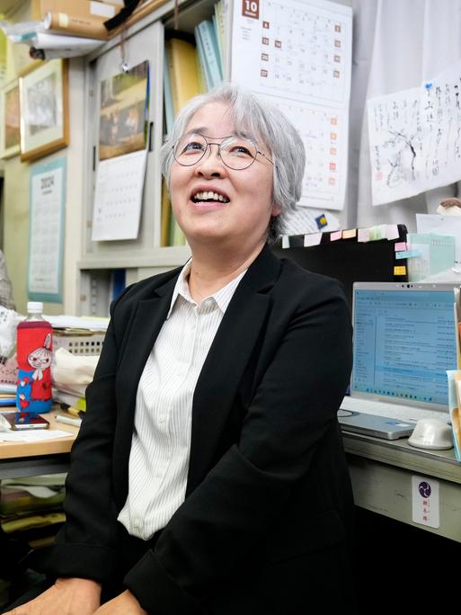 Nihon Hidanjyo sitzt auf einem Bürostuhl und lächelt. Im Hintergrund ist ein Fotoapparat auf sie gerichtet. Sie hat weiße kurze Haare und trägt eine Brille.