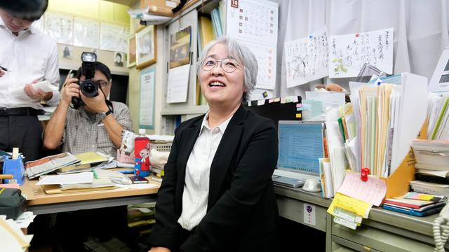 Nihon Hidanjyo sitzt auf einem Bürostuhl und lächelt. Im Hintergrund ist ein Fotoapparat auf sie gerichtet. Sie hat weiße kurze Haare und trägt eine Brille.