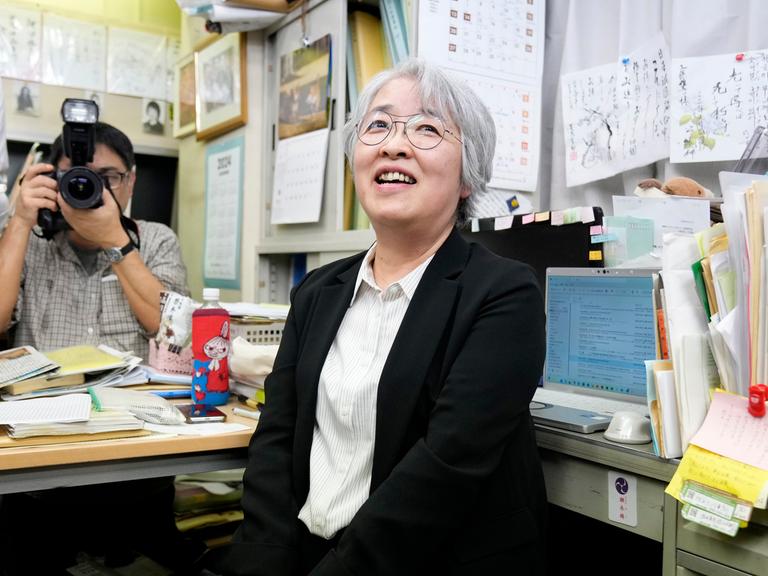 Nihon Hidanjyo sitzt auf einem Bürostuhl und lächelt. Im Hintergrund ist ein Fotoapparat auf sie gerichtet. Sie hat weiße kurze Haare und trägt eine Brille.