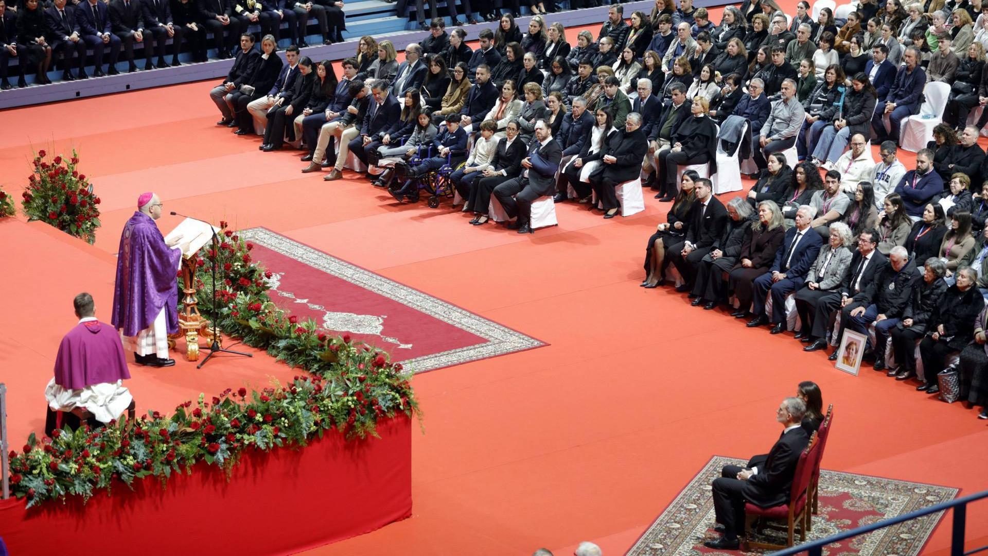 Viele Menschen sitzen in schwarzer Kleidung auf Bänken. Ein Pastor predigt auf einer Kanzel. Rechts sitzen der spanische König und seine Ehefrau.