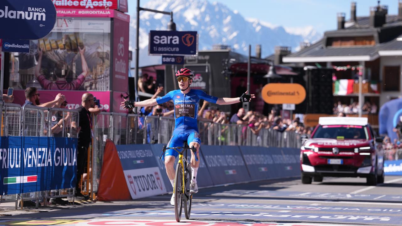 Jonas Vingegaard fährt über die Ziellinie beim Giro d'Italia. Er trägt ein blaues Trikot und einen roten Helm. Hinter ihm fährt ein rotes Auto. 