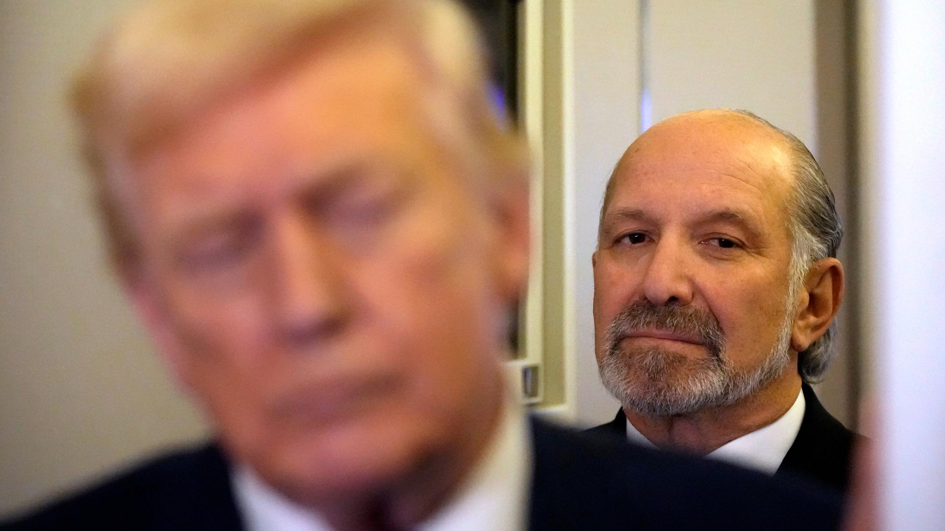 Im Vordergrund des Bildes ist unscharf Trump zu sehen. Im Hintergrund Howard Lutnick, der mit ernster Miene zuhört.