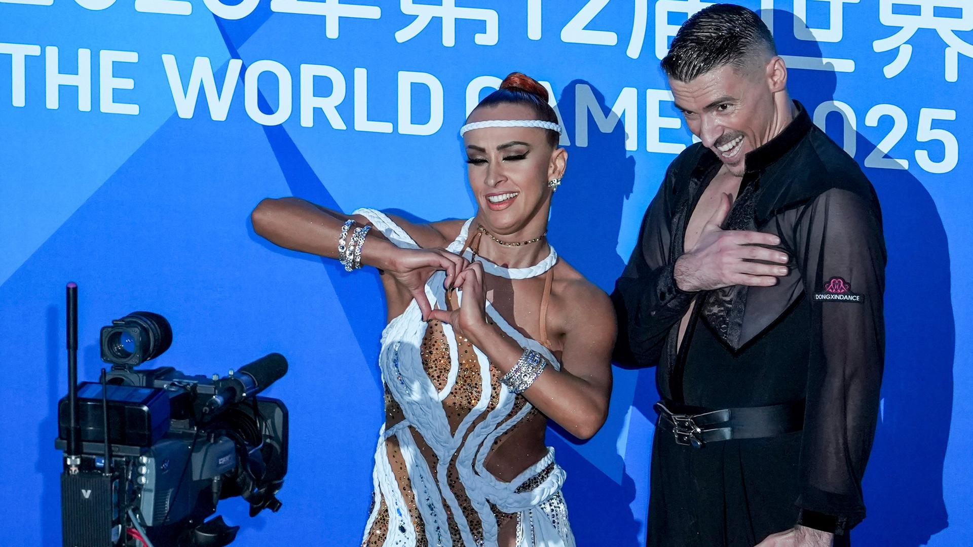 Marius Balan (R) und Khrystyna Moshenska am Rande des Latin-Final-Tanzes bei den World Games 2025 in Chengdu. Moshenska zeigt ein mit den Händen geformtes Herz in die eine Kamera