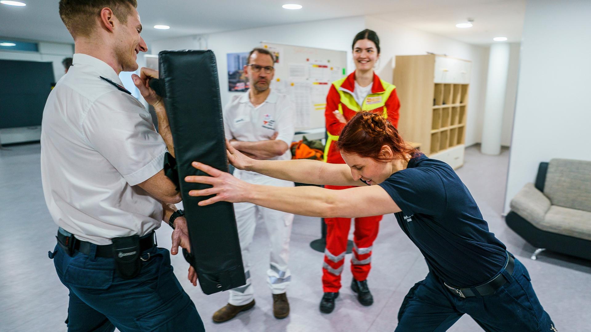 Eine Frau in Uniform stemmt sich mit aller Kraft gegen ein schwarzes Kissen, das ein Mann in Uniform vor sich hält. Im Hintergrund stehen ein weiterer uniformierter Mann und eine weitere uniformierte Frau.