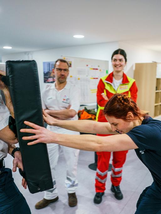 Eine Frau in Uniform stemmt sich mit aller Kraft gegen ein schwarzes Kissen, das ein Mann in Uniform vor sich hält. Im Hintergrund stehen ein weiterer uniformierter Mann und eine weitere uniformierte Frau.