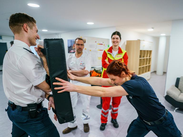 Eine Frau in Uniform stemmt sich mit aller Kraft gegen ein schwarzes Kissen, das ein Mann in Uniform vor sich hält. Im Hintergrund stehen ein weiterer uniformierter Mann und eine weitere uniformierte Frau.