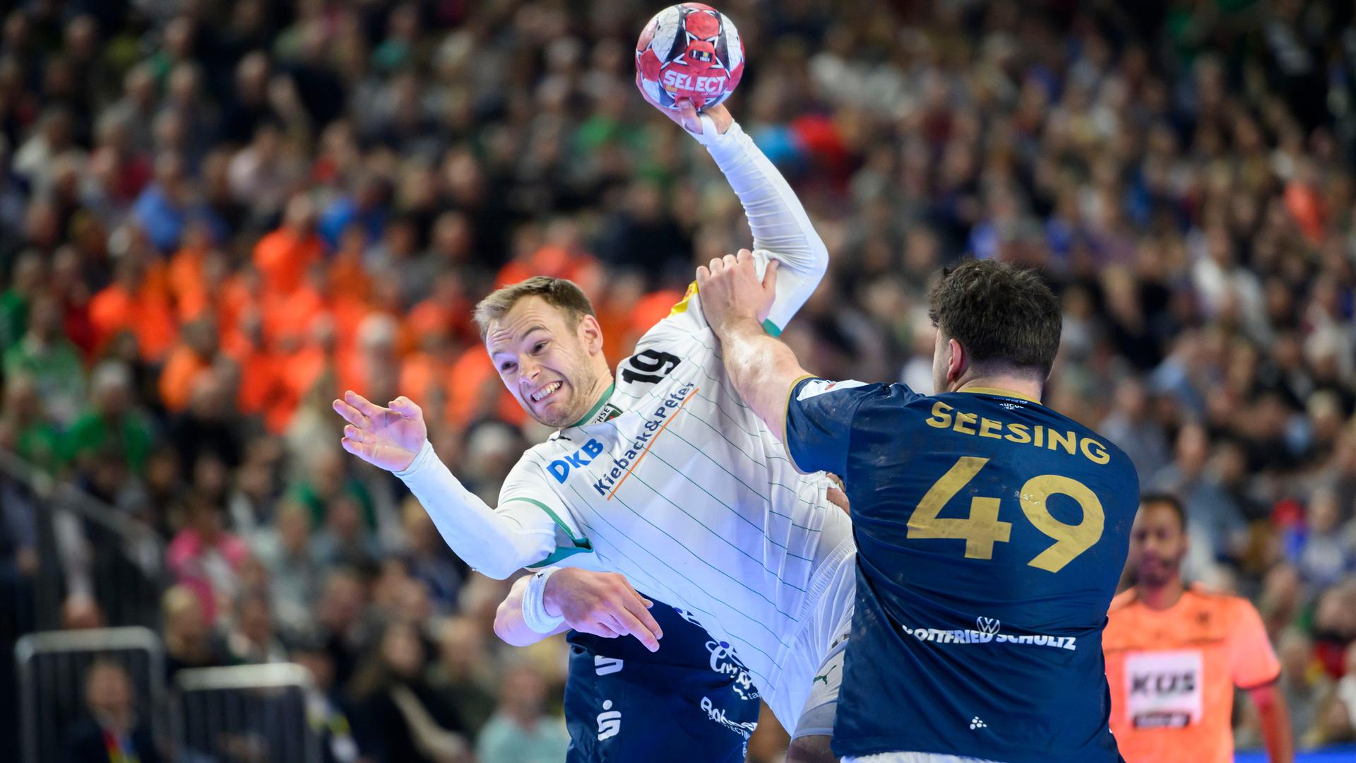 Gidsel im weißen Trikot wirft gerade, Seesing im dunkelblauen Trikot sowie ein weiterer Spieler des Bergischen HC, der von Gidsel verdeckt ist, wollen ihn daran hindern. Dahinter unscharf ein Schiedsrichter und eine vollbesetzte Tribüne.