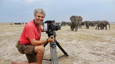 Porträt von Tierfilmer Jens Westphalen mit seiner Kamera. Im Hintergrund ist eine Herde Elefanten in Botswana zu sehen Porträt von Tierfilmer Jens Westphalen mit seiner Kamera. Im Hintergrund ist eine Herde Elefanten in Botswana zu sehen