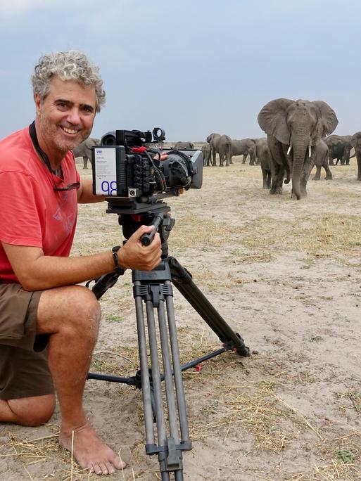 Porträt von Tierfilmer Jens Westphalen mit seiner Kamera. Im Hintergrund ist eine Herde Elefanten in Botswana zu sehen 