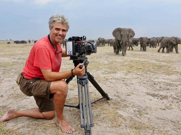 Porträt von Tierfilmer Jens Westphalen mit seiner Kamera. Im Hintergrund ist eine Herde Elefanten in Botswana zu sehen 