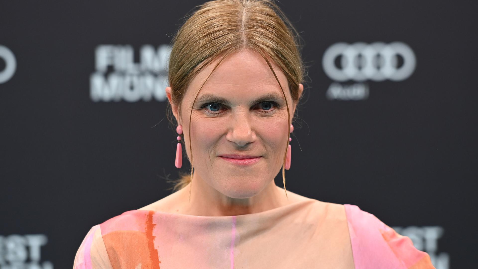 Die Schauspielerin Fritzi Haberlandt schaut in die Kamera. Sie hat blondes zusammengebundenes Haar und rosa, längliche Ohrringe. 