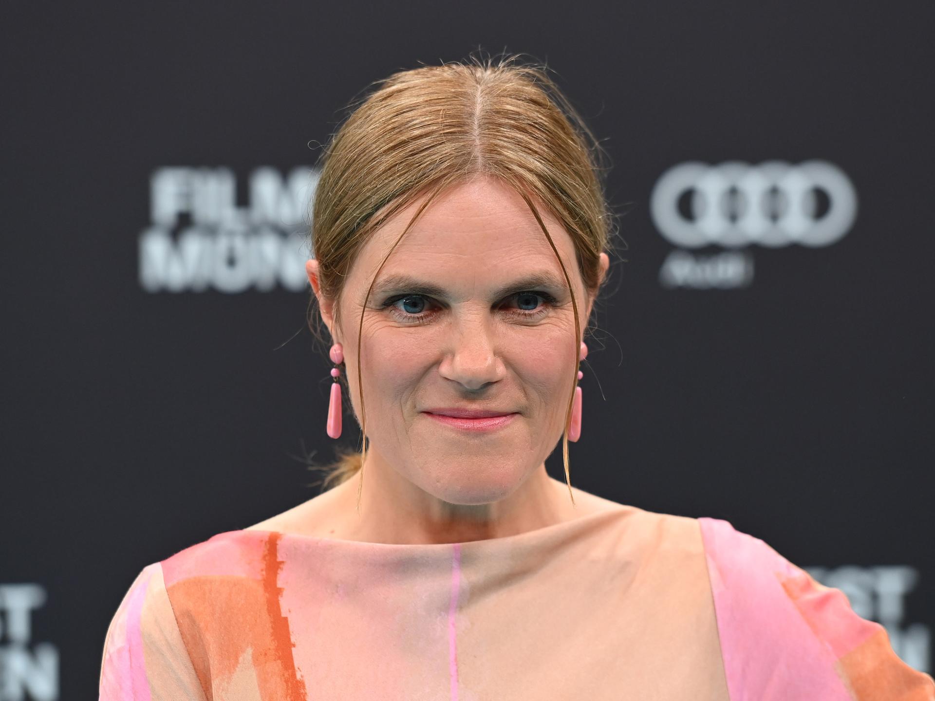 Die Schauspielerin Fritzi Haberlandt schaut in die Kamera. Sie hat blondes zusammengebundenes Haar und rosa, längliche Ohrringe. 