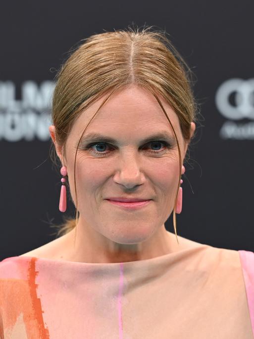 Die Schauspielerin Fritzi Haberlandt schaut in die Kamera. Sie hat blondes zusammengebundenes Haar und rosa, längliche Ohrringe. 
