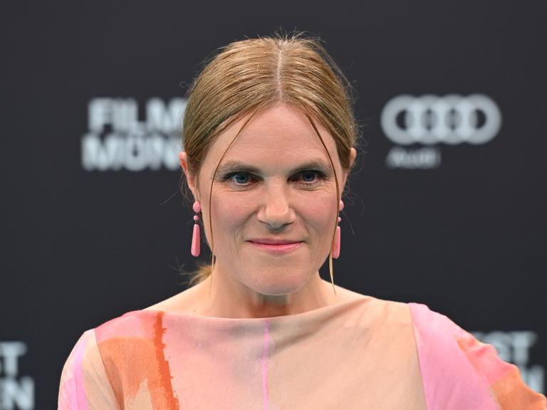 Die Schauspielerin Fritzi Haberlandt schaut in die Kamera. Sie hat blondes zusammengebundenes Haar und rosa, längliche Ohrringe. 