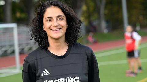 Die Fußballerin Tugba Tekkal, Ex-Profi der Frauen-Bundesliga, steht auf einem Fußballplatz. Sie hat lockige, dunkle Haare trägt ein schwarzes Fußballshirt mit der Aufschrift "Scoring Girls".