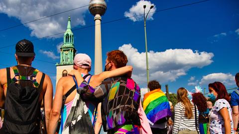 In einer Reihe stehen mehrere Demonstrierende beim CSD in bunter Kleidung, eine Person legt einer anderen den Arm um die Schulter. In einer Reihe stehen mehrere Demonstrierende beim CSD in bunter Kleidung, eine Person legt einer anderen den Arm um die Schulter.