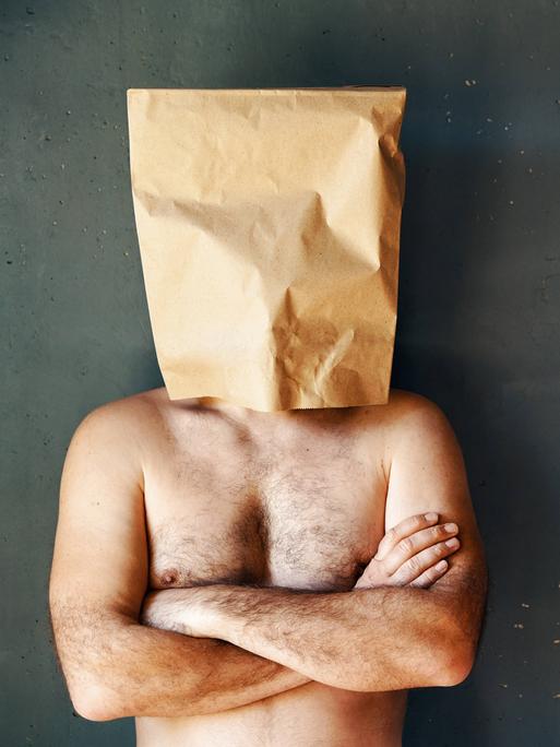 Ein Mann mit freiem Oberkörper hat eine Papiertüte über den Kopf gestülpt. Seine Arme sind verschränkt. Ein Mann mit freiem Oberkörper hat eine Papiertüte über den Kopf gestülpt. Seine Arme sind verschränkt.