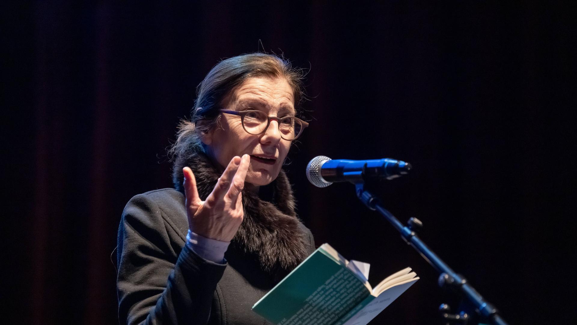 Die Schriftstellerin Ursula Krechel steht bei einer Lesung in der Berliner Volksbühne auf dem Podium an einem Mikrofon und hält ein aufgeklapptes Buch in der Hand.