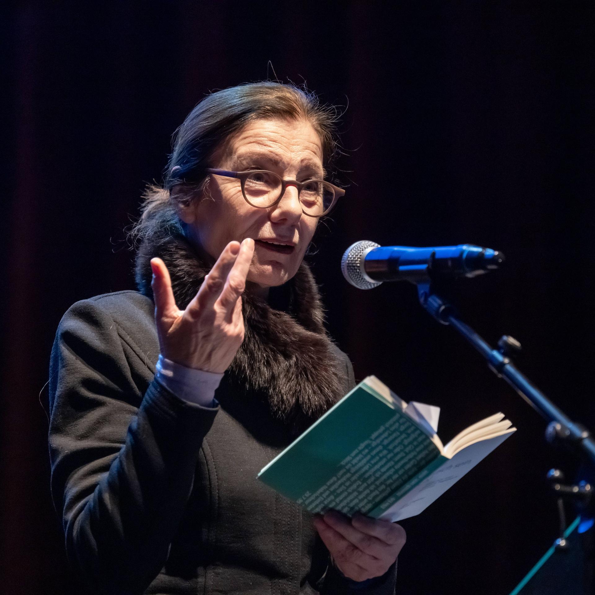 Die Schriftstellerin Ursula Krechel steht bei einer Lesung in der Berliner Volksbühne auf dem Podium an einem Mikrofon und hält ein aufgeklapptes Buch in der Hand.