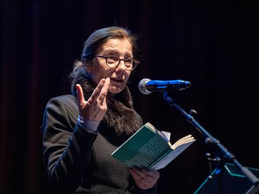 Die Schriftstellerin Ursula Krechel steht bei einer Lesung in der Berliner Volksbühne auf dem Podium an einem Mikrofon und hält ein aufgeklapptes Buch in der Hand.
