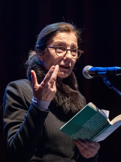 Die Schriftstellerin Ursula Krechel steht bei einer Lesung in der Berliner Volksbühne auf dem Podium an einem Mikrofon und hält ein aufgeklapptes Buch in der Hand.