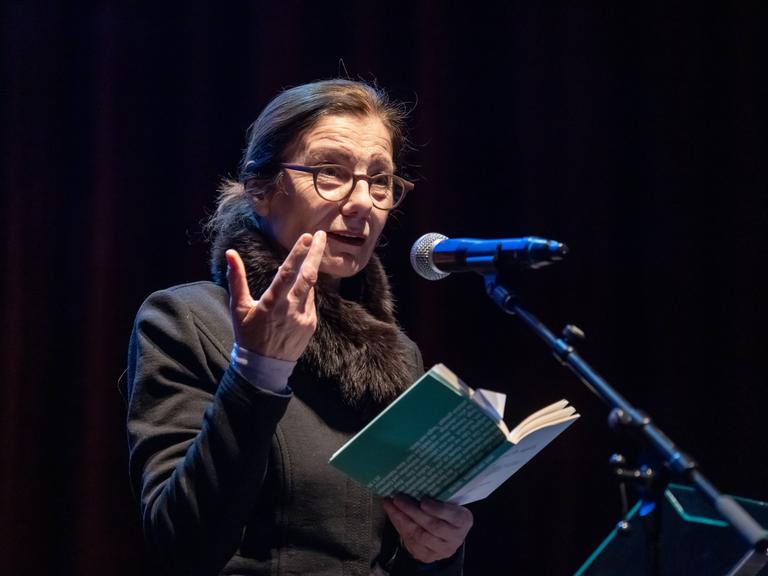 Die Schriftstellerin Ursula Krechel steht bei einer Lesung in der Berliner Volksbühne auf dem Podium an einem Mikrofon und hält ein aufgeklapptes Buch in der Hand.