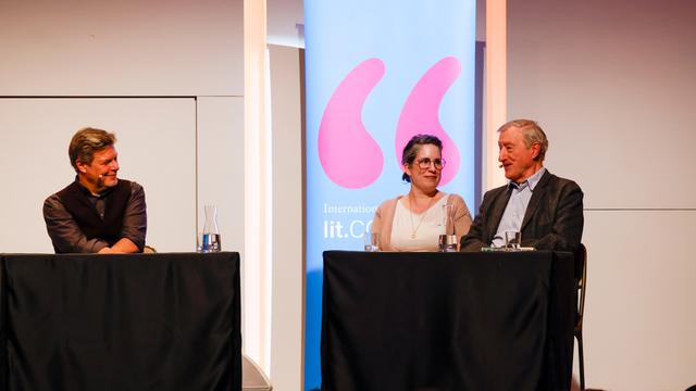 Auf einer Bühne sitzen drei Menschen vor Mikrofonen. Vor ihnen sitzen viele Menschen im Publikum.